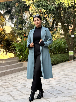 Woman in a long light blue coat standing outdoors with greenery in the background