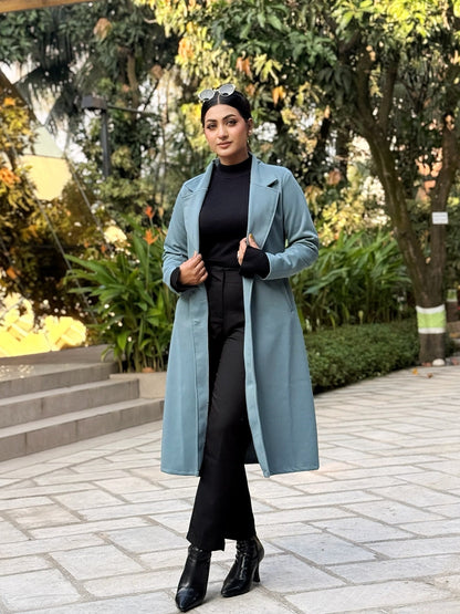 Woman in a long light blue coat standing outdoors with greenery in the background