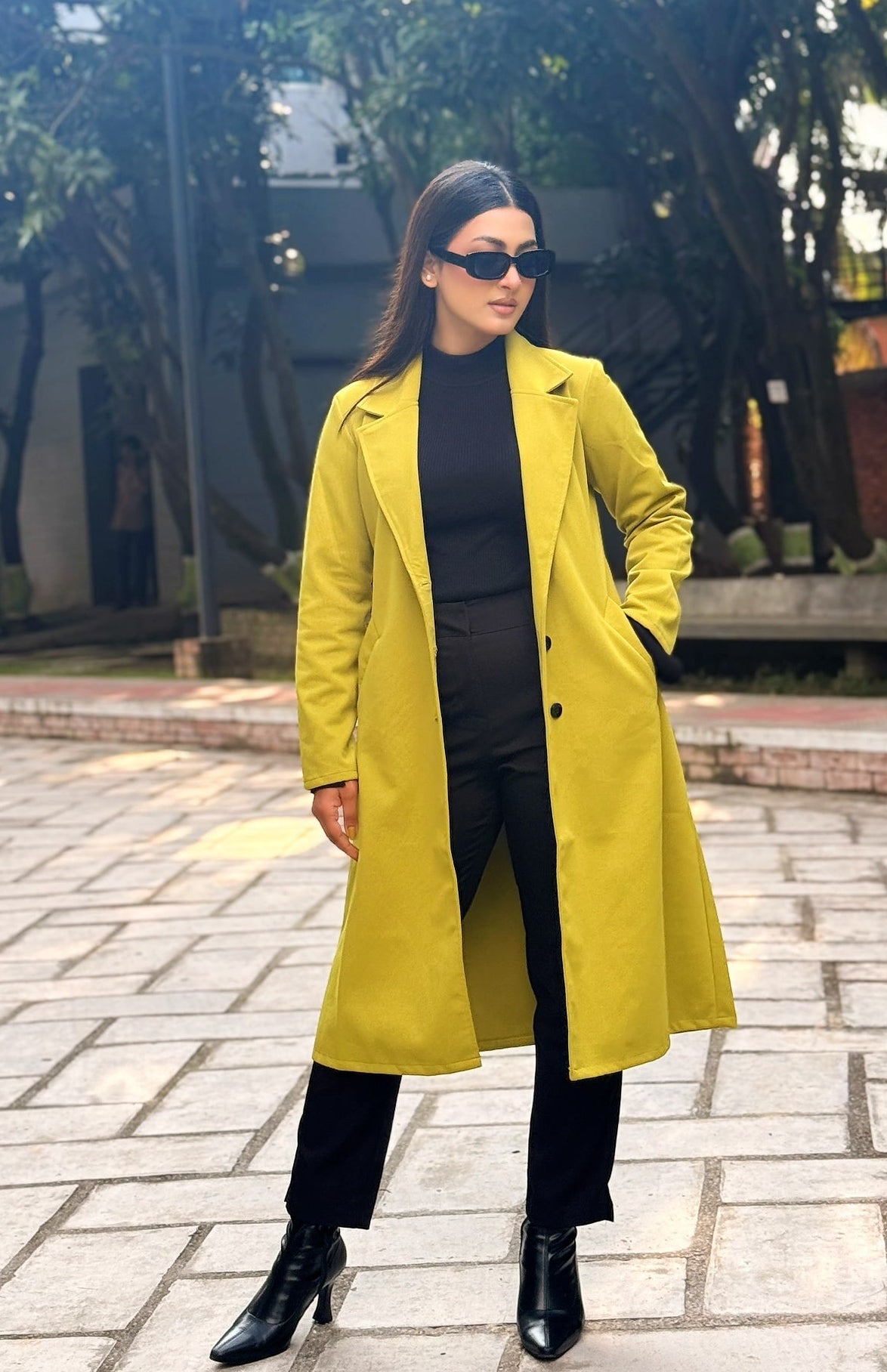 Woman wearing a yellow coat and black outfit standing outdoors on a paved area with trees in the background.
