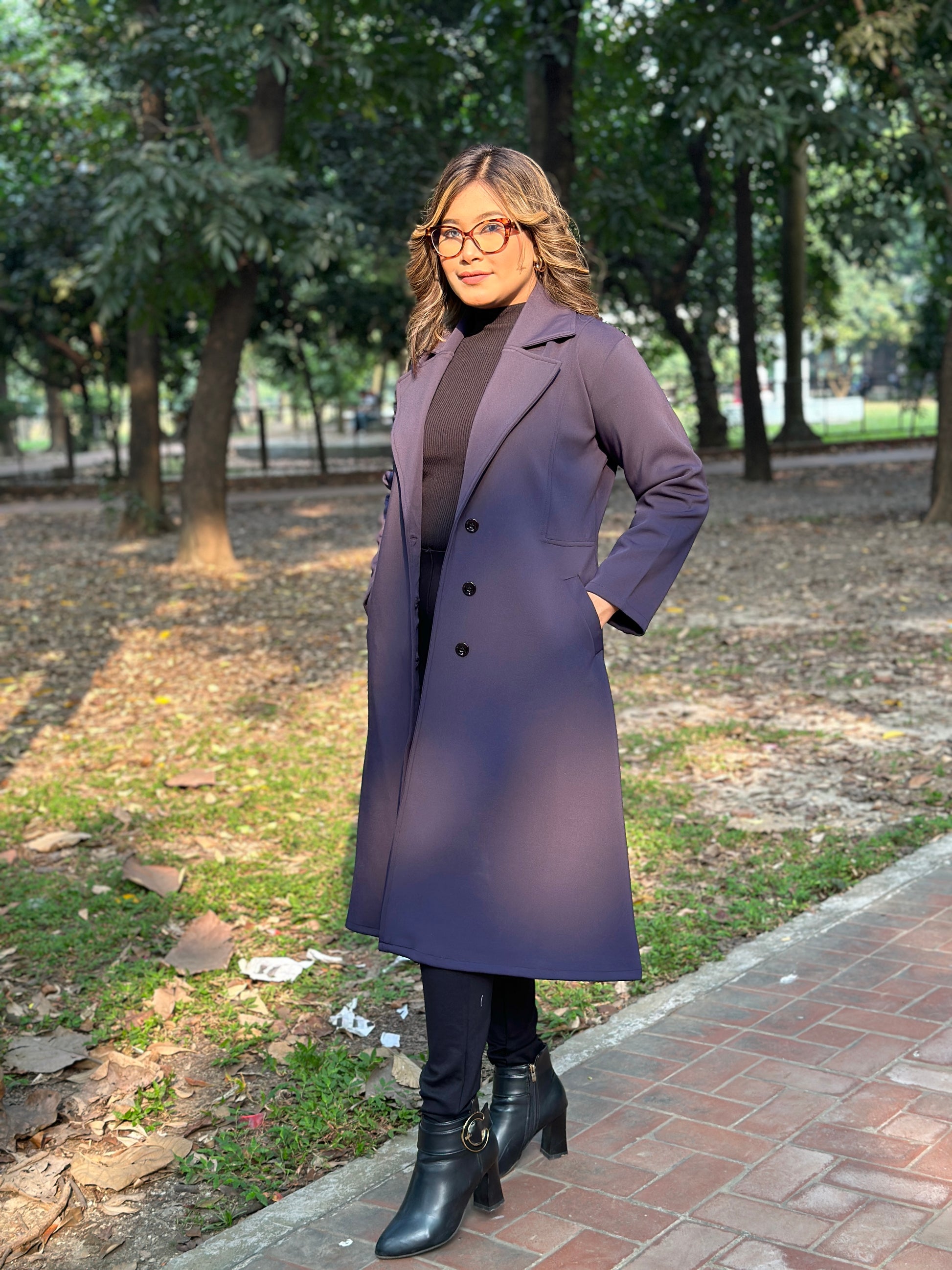 Woman wearing a long blueish black overcoat standing on a sidewalk with trees in the background