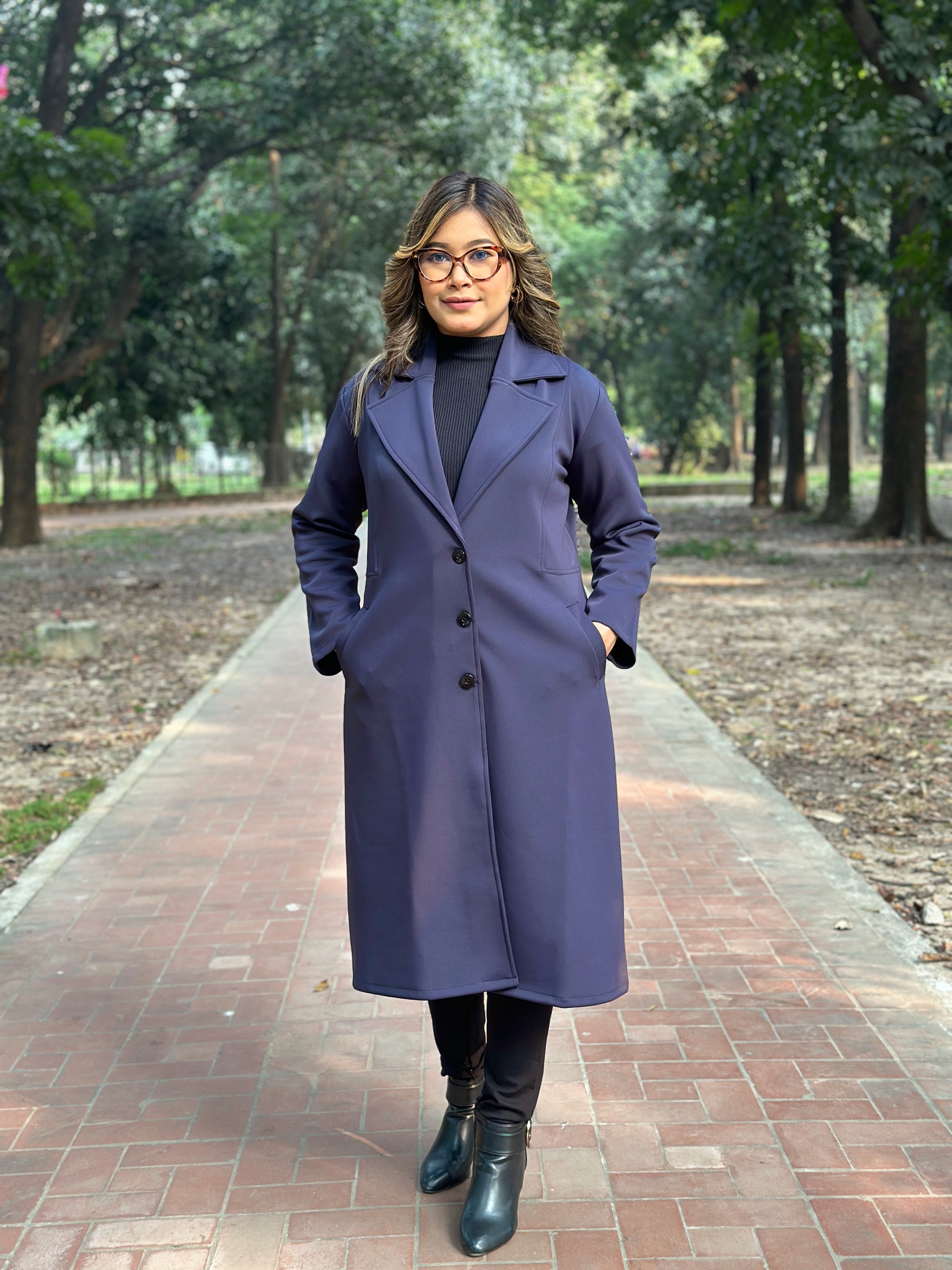 Woman wearing a long blueish black coat standing on a brick path with trees in the background