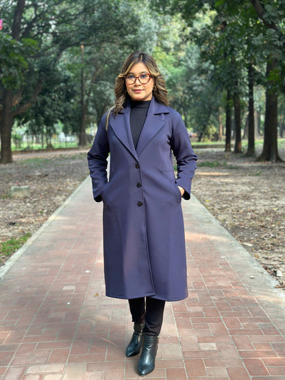 Woman wearing a long blueish black coat standing on a brick path with trees in the background