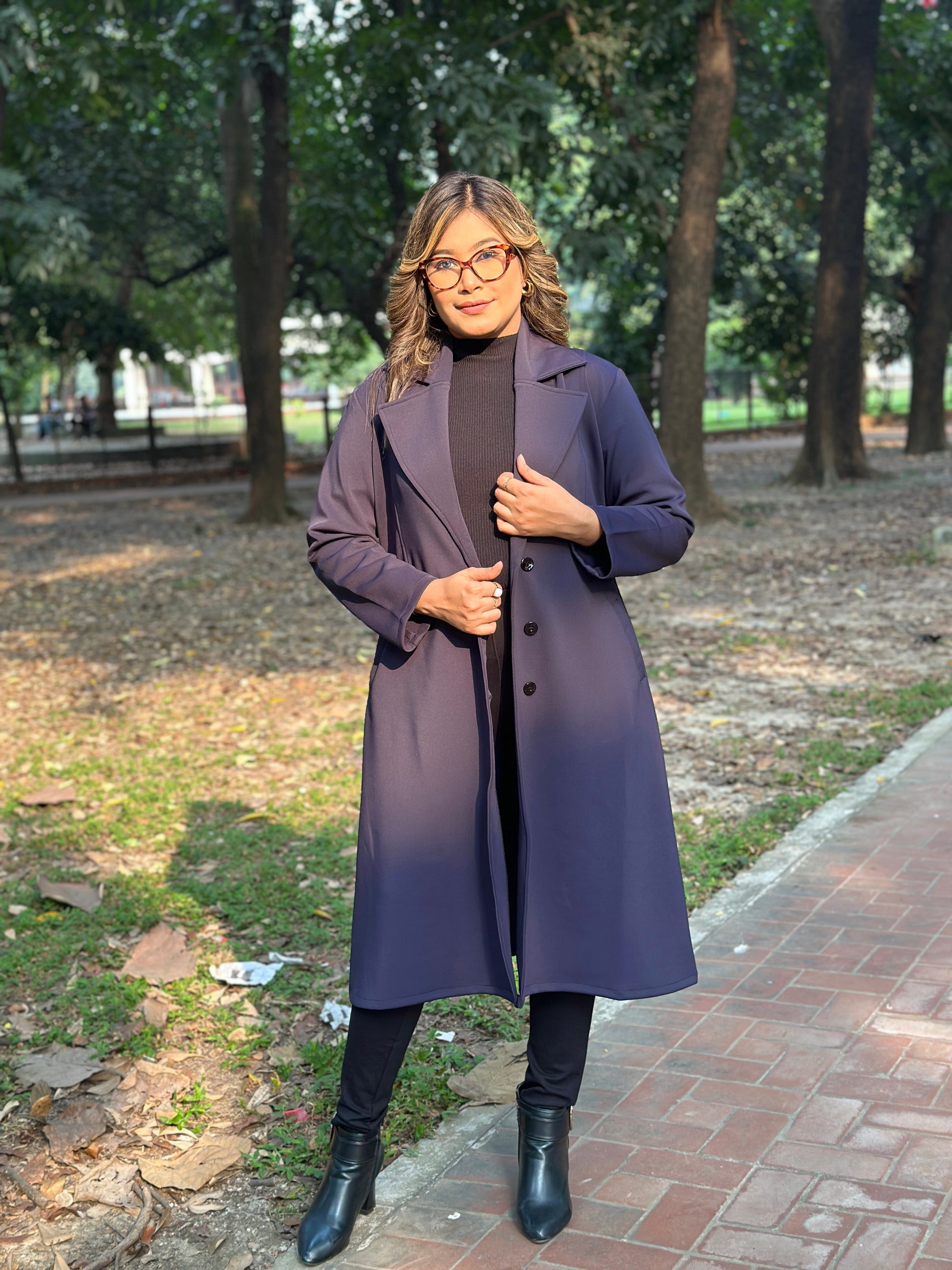 Person wearing a long blueish black overcoat standing on a sidewalk with trees in the background