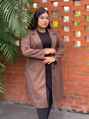 Woman wearing a brown coat standing in front of a brick wall with plants.