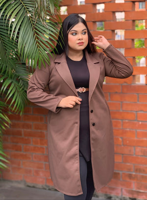 Woman wearing a brown coat standing against a brick wall with plants.