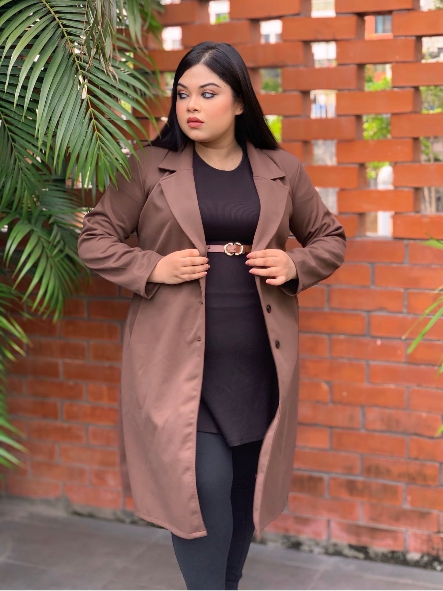 Woman wearing a brown coat standing against a brick wall with plants.