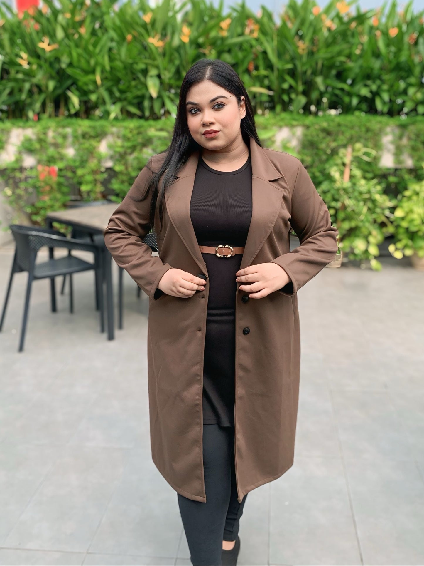 Woman wearing a brown coat standing outdoors with greenery in the background