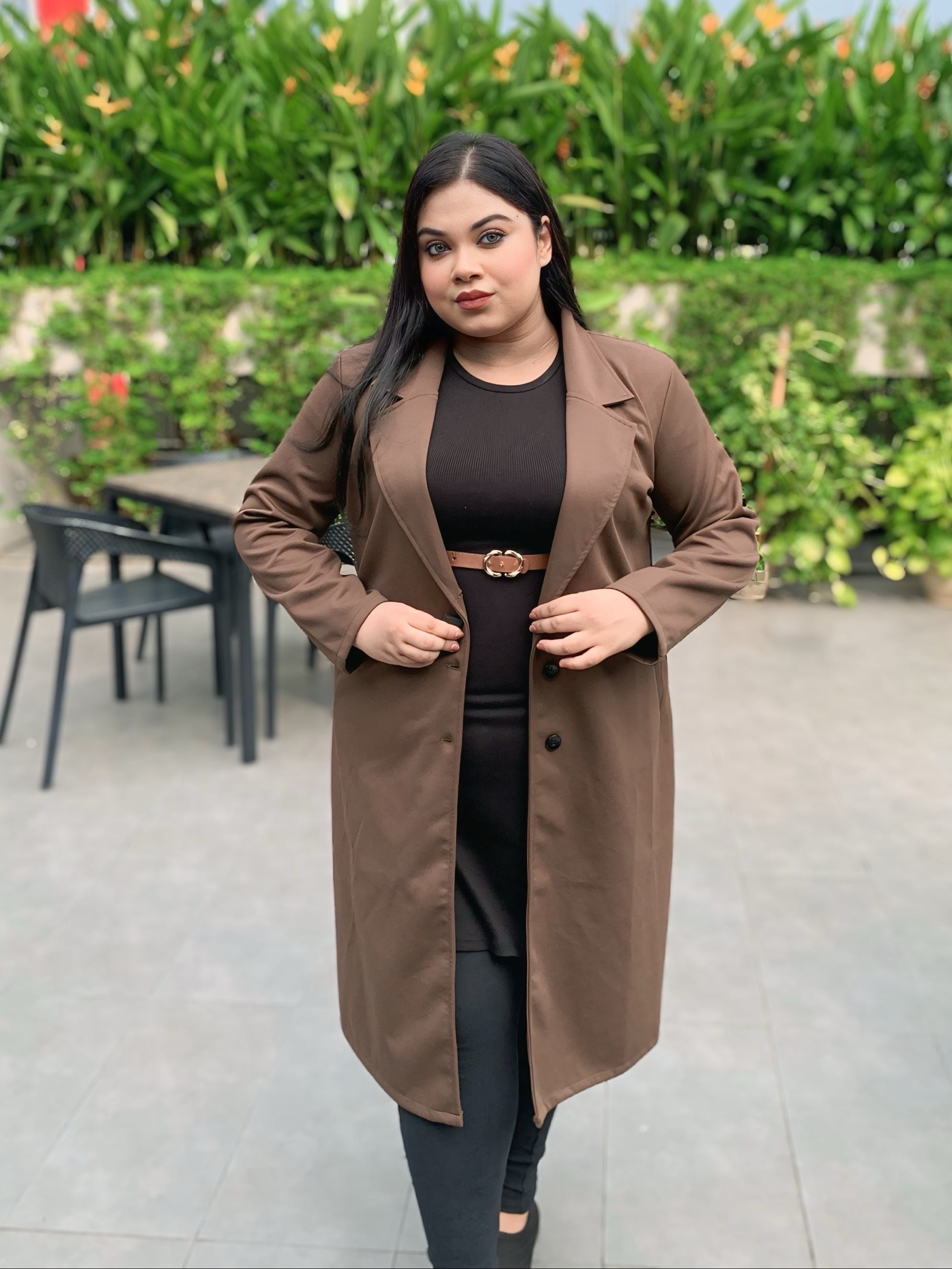 Woman wearing a brown coat standing outdoors with greenery in the background