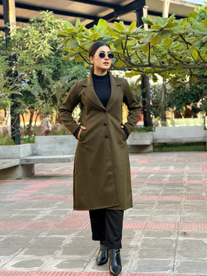 Person wearing an olive green coat in an outdoor setting with trees and a building in the background.