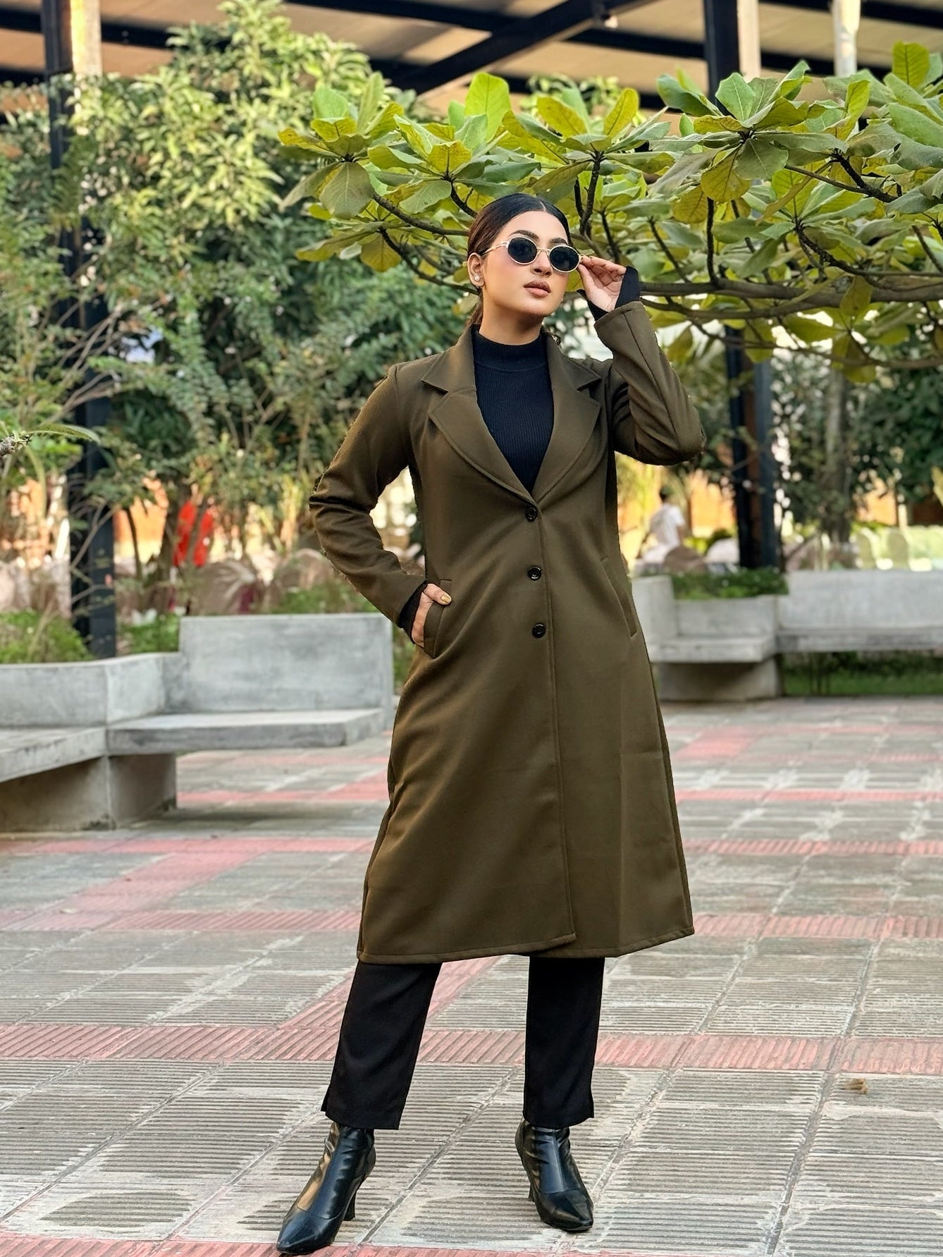 Person wearing a long brown coat in an outdoor setting with greenery and benches.