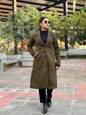 Person wearing a long olive green coat in an outdoor setting with trees and benches.