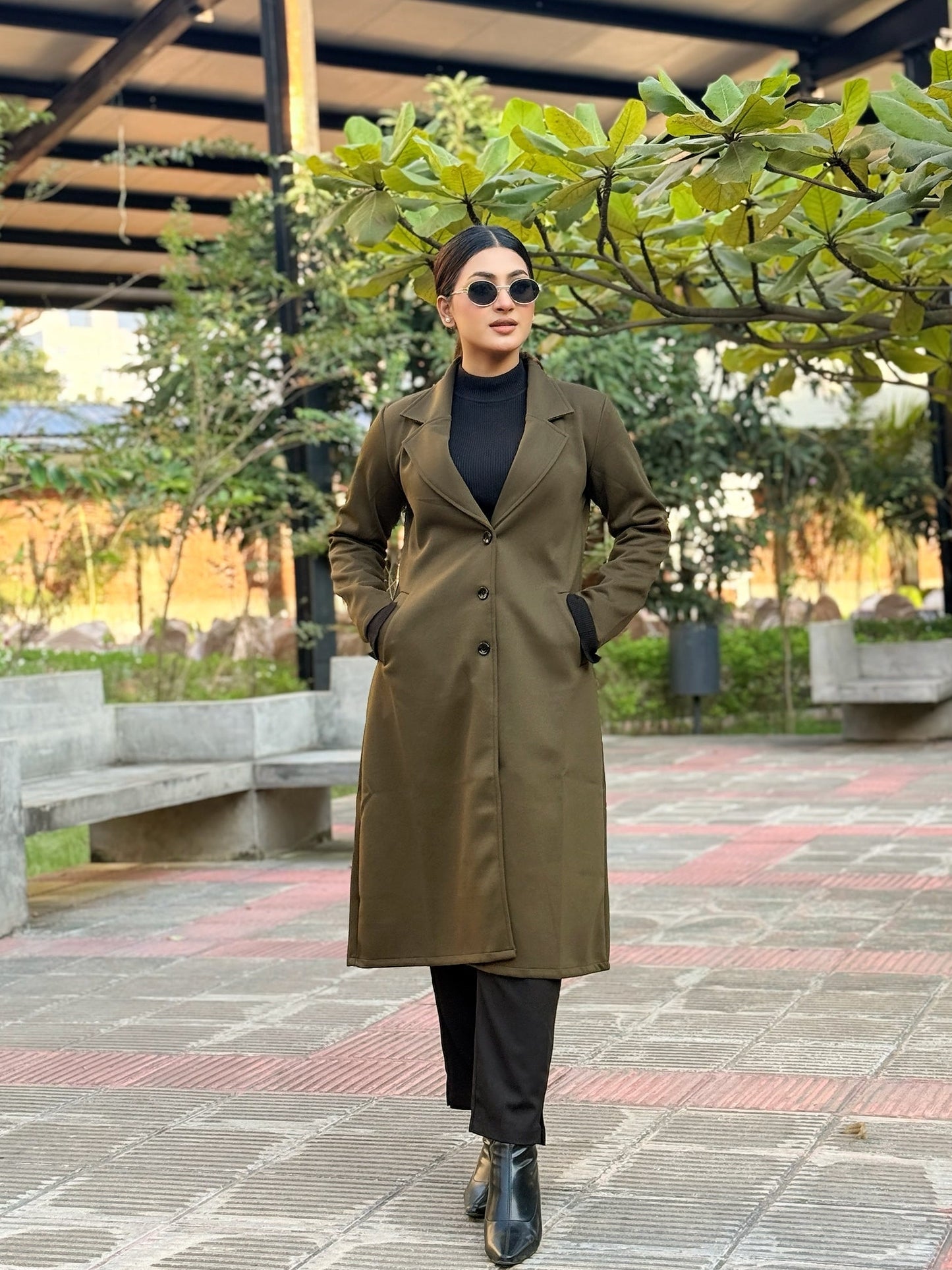 Person wearing a long green coat and black pants standing outdoors on a paved area with trees and benches in the background.