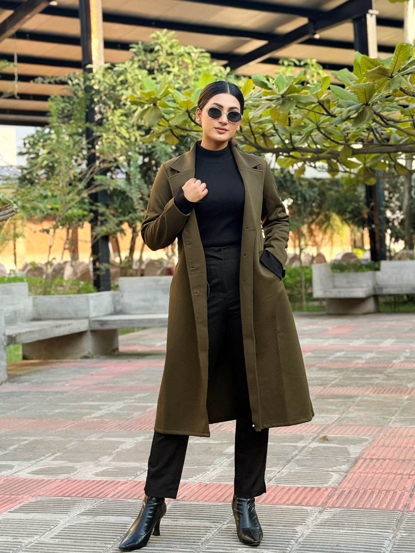 Woman in a long brown coat standing outdoors with trees and benches in the background