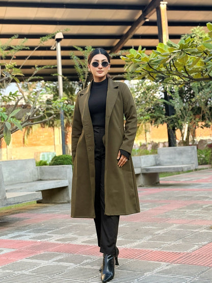 Woman in a green coat standing outdoors on a paved area with trees and benches in the background.