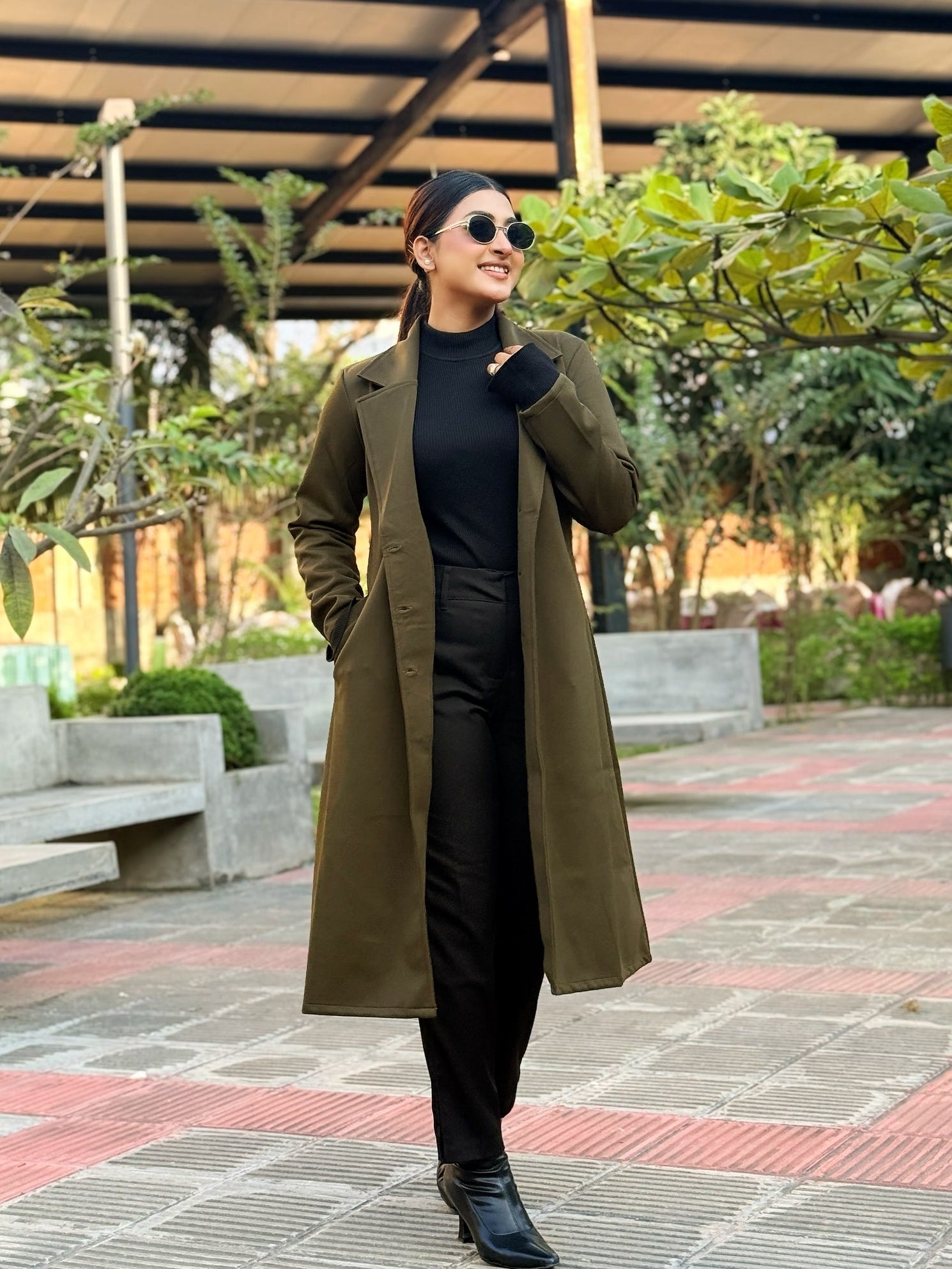 Woman in a long green coat standing outdoors with trees and a building in the background