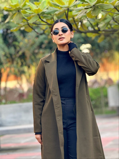 Woman wearing a long olive green coat and sunglasses in an outdoor setting with trees and benches.