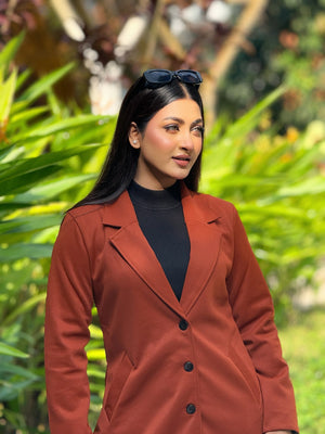Woman wearing a brown coat and black top outdoors with greenery in the background