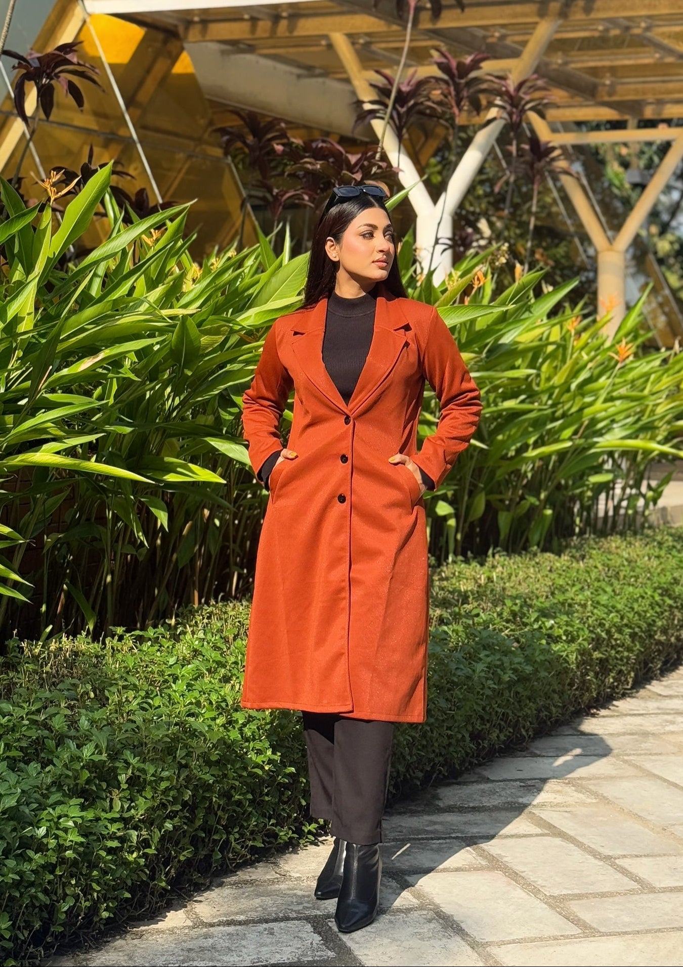 Woman in an orange coat standing in a garden with greenery and a building in the background