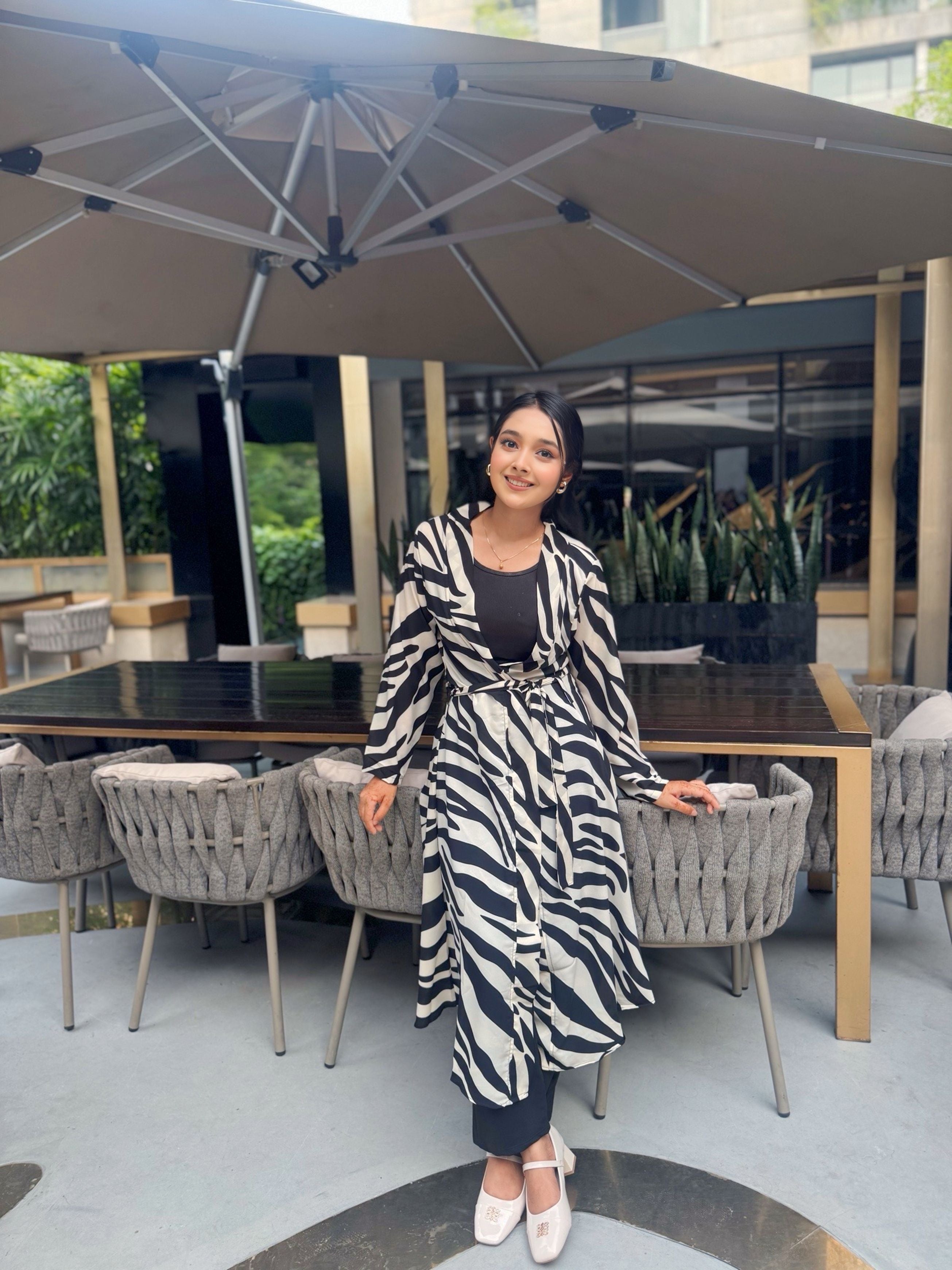 Woman in a zebra print long shrug sitting at an outdoor dining table with a large umbrella.