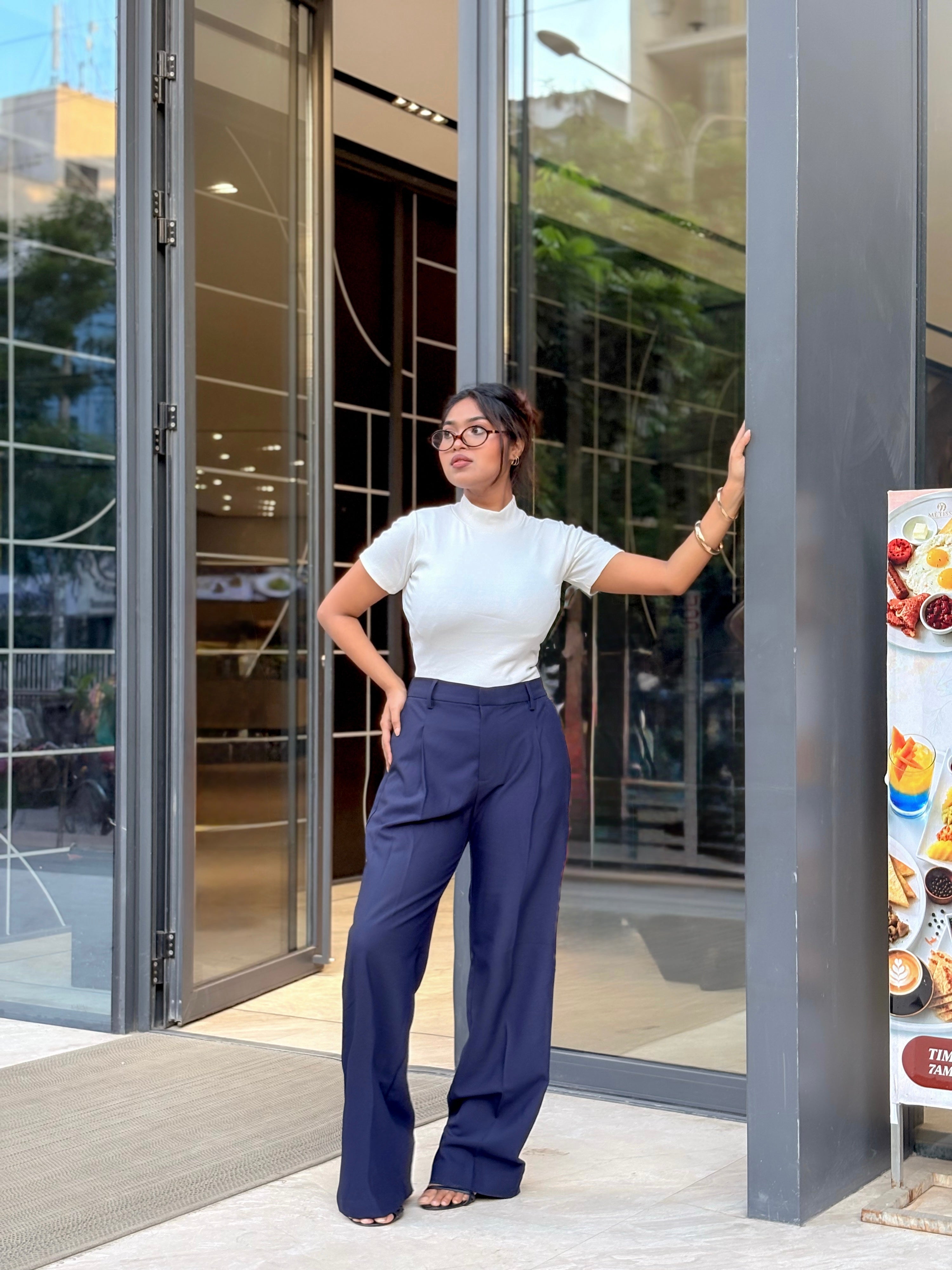 A woman wearing a white short-sleeve mock neck top and navy blue wide-leg trousers stands in front of a modern glass building entrance.