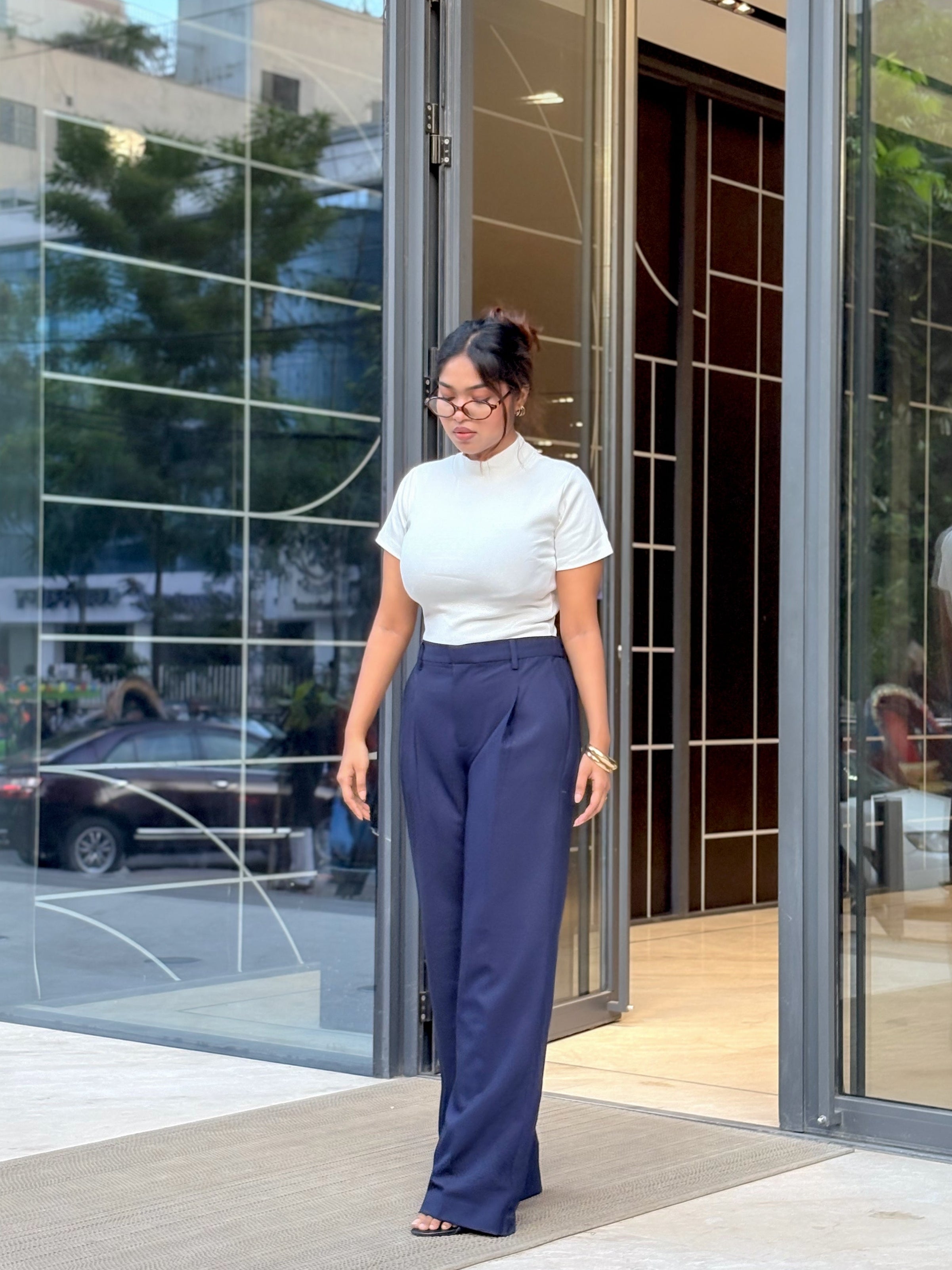 A woman wearing a white short-sleeve mock neck top and navy blue wide-leg trousers walks out of a modern glass building entrance.