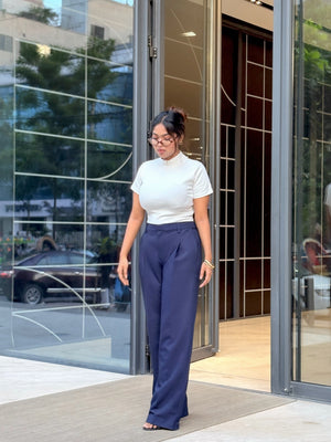 A woman wearing a white short-sleeve mock neck top and navy blue wide-leg trousers walks out of a modern glass building entrance.
