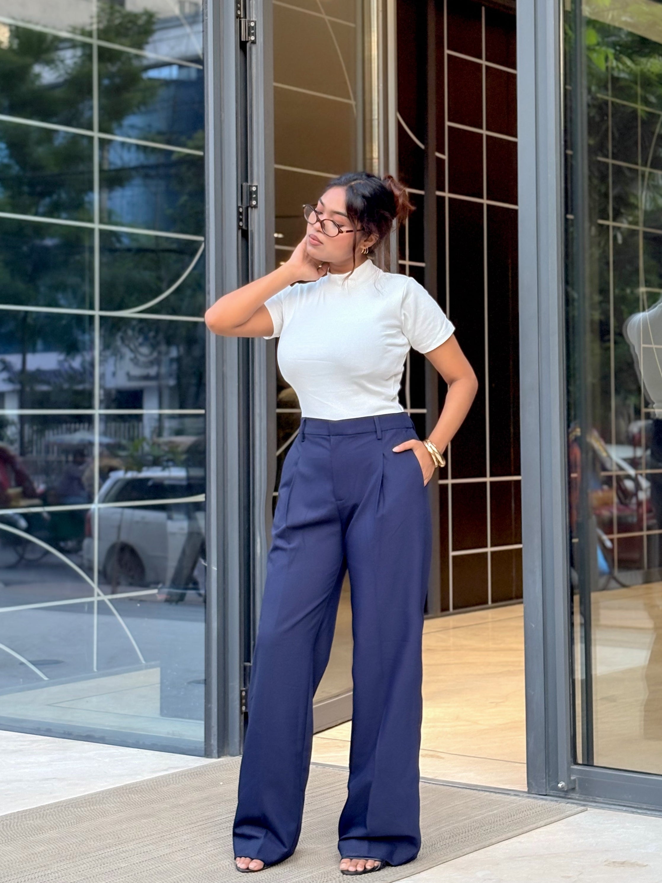 A woman wearing a white short-sleeve top and navy blue wide-leg trousers stands in front of a modern glass storefront.