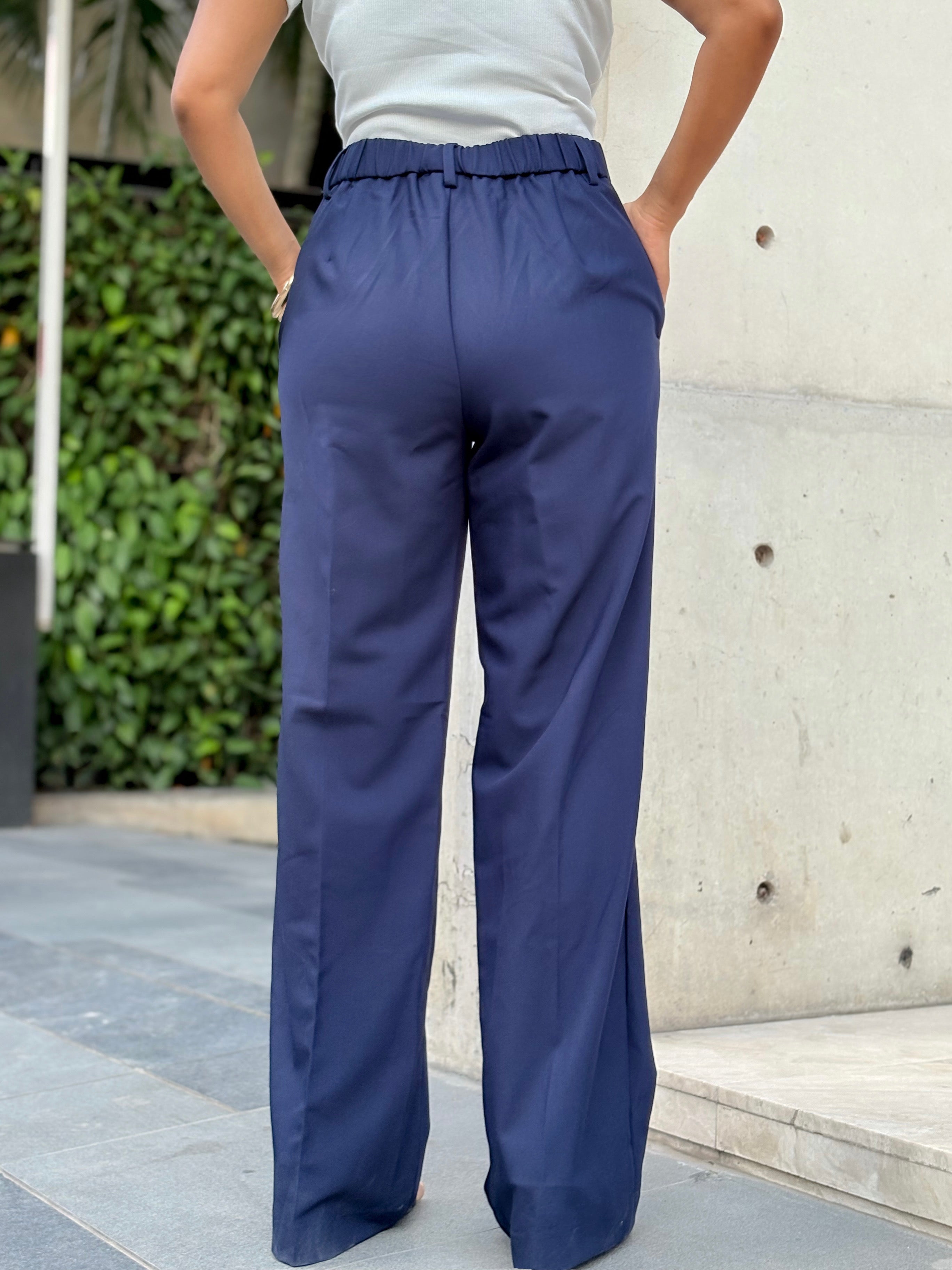 A person shown from the back wearing navy wide-leg trousers with an elastic waistband, standing outdoors against a light concrete wall and greenery.