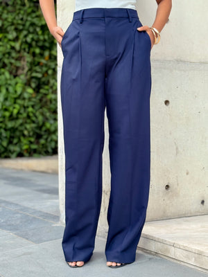 A person wearing high-waisted, wide-leg navy blue trousers with front pleats, standing outdoors against a light concrete wall.