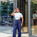 A woman stands in front of a modern glass building entrance wearing a white short-sleeve mock neck top and navy blue wide-leg trousers.