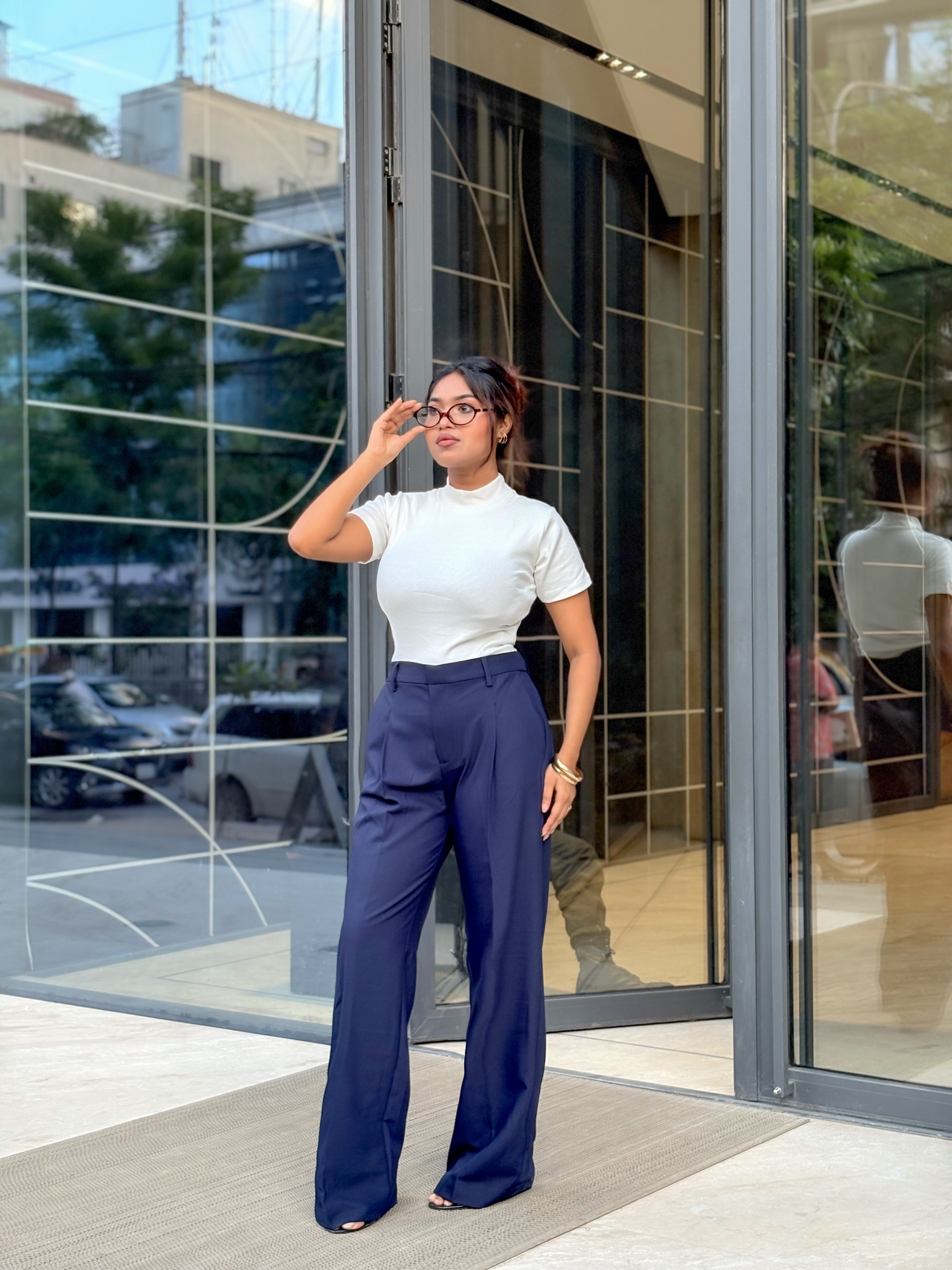 A woman stands in front of a modern glass building entrance wearing a white short-sleeve mock neck top and navy blue wide-leg trousers.