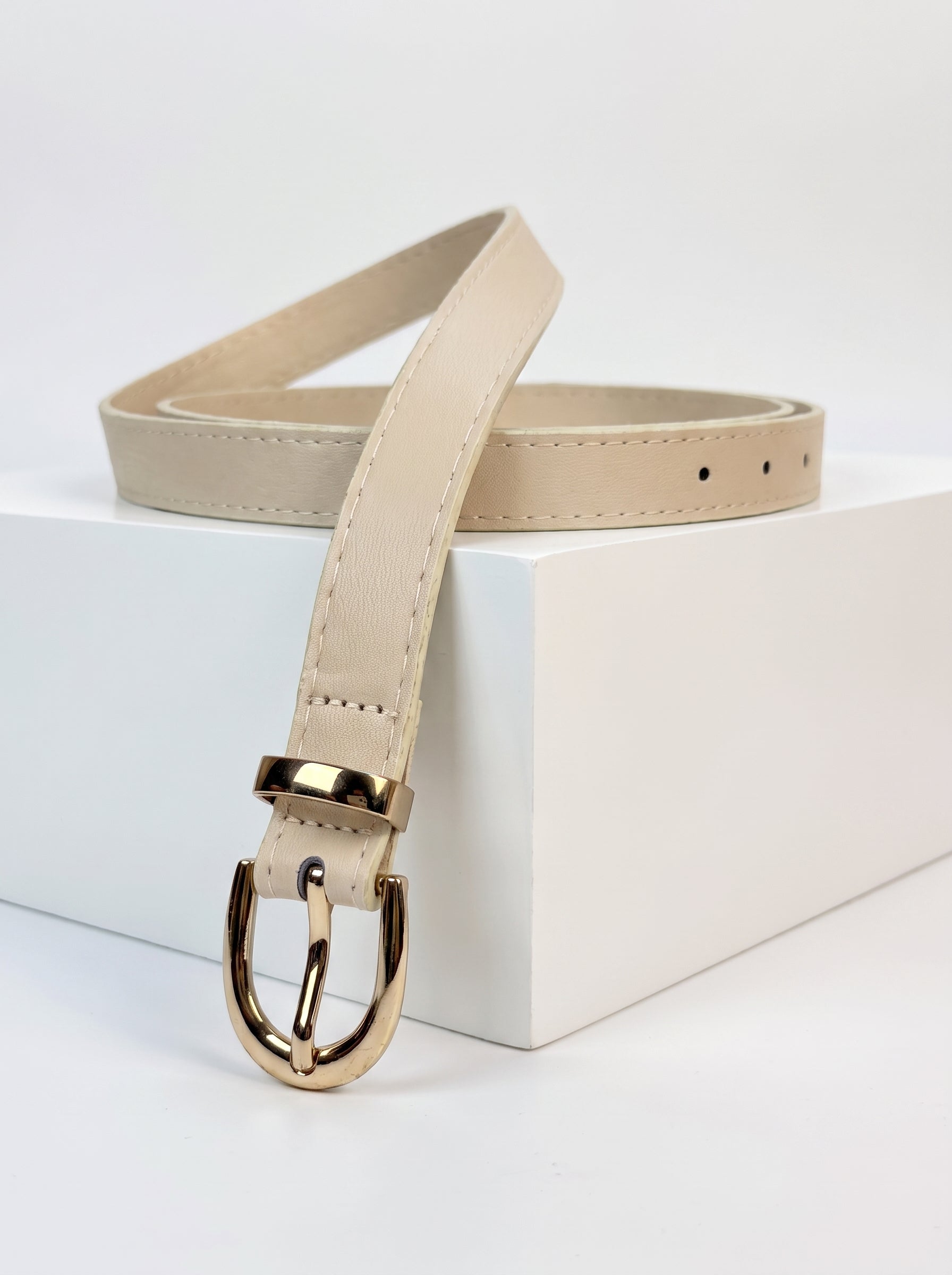 A light beige leather belt with a polished gold-tone metal buckle, displayed on a white geometric block against a clean white background.