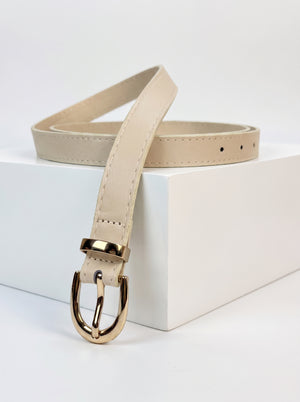 A light beige leather belt with a polished gold-tone metal buckle, displayed on a white geometric block against a clean white background.