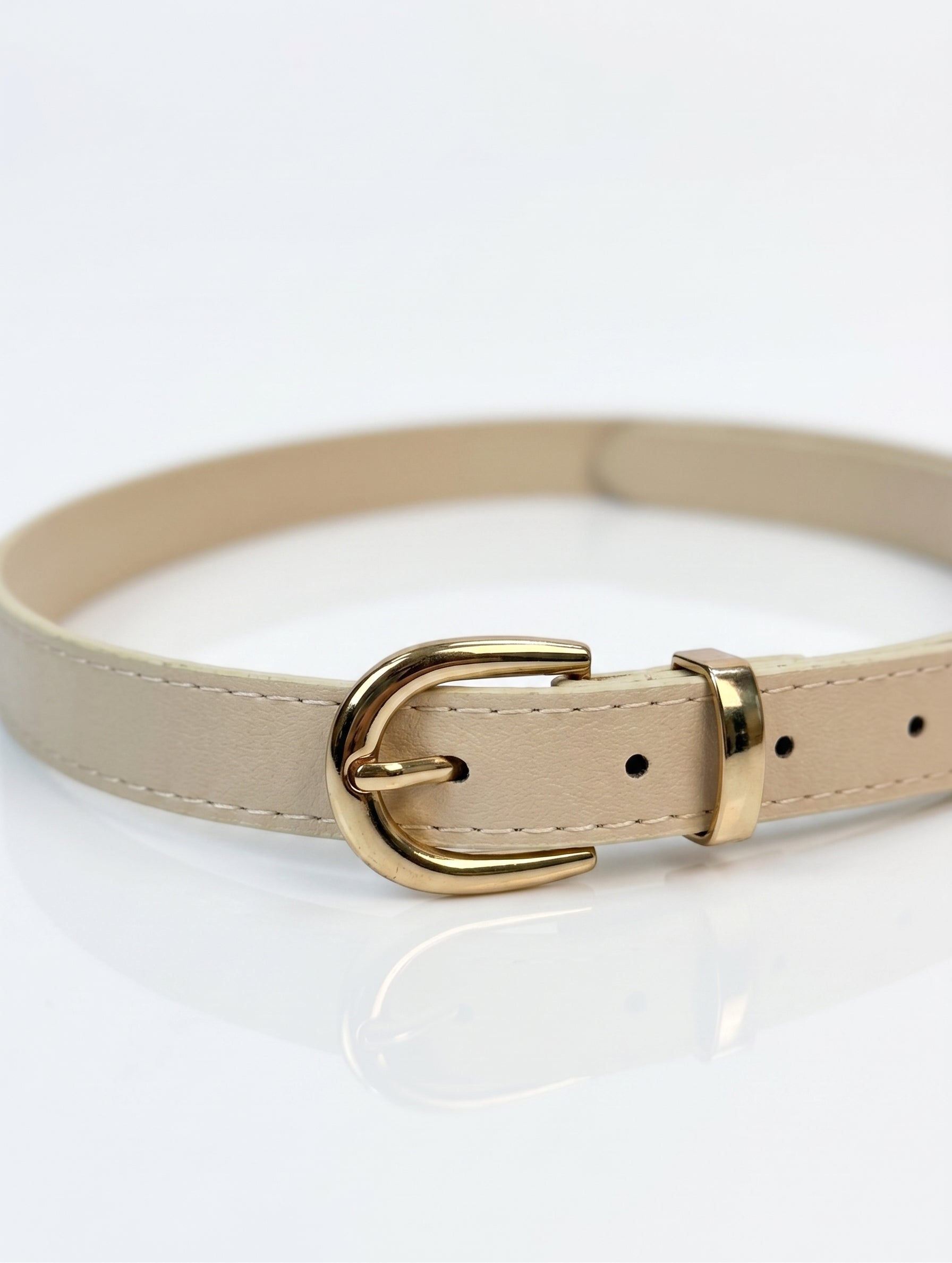 A beige faux leather belt with a polished gold-tone metal buckle, shown coiled on a reflective white surface.