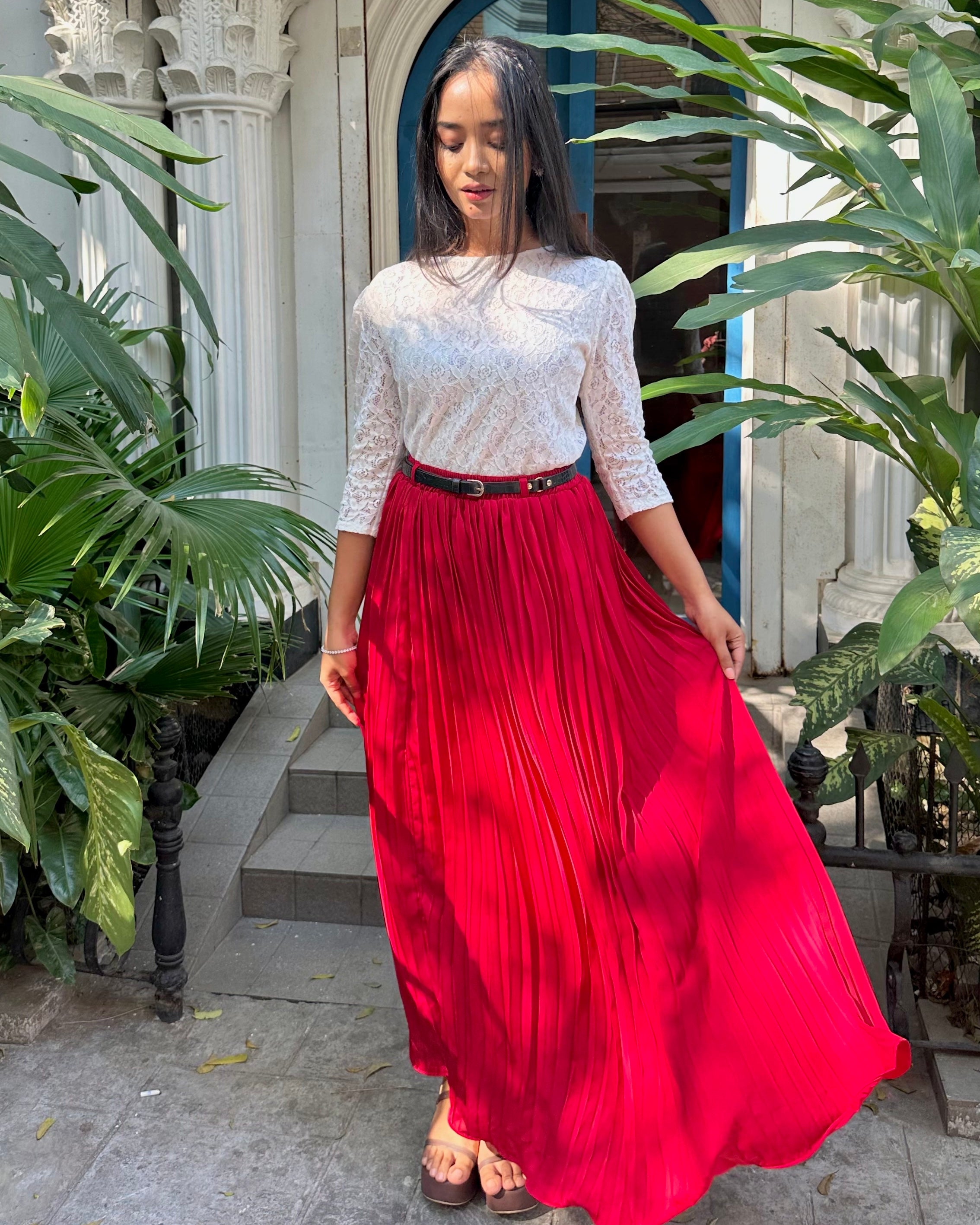 Woman wearing a white top and red pleated skirt standing in front of a building with plants.