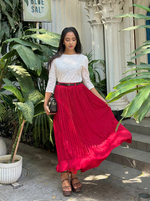 Woman in a white top and red skirt standing outdoors with plants and architectural elements in the background.