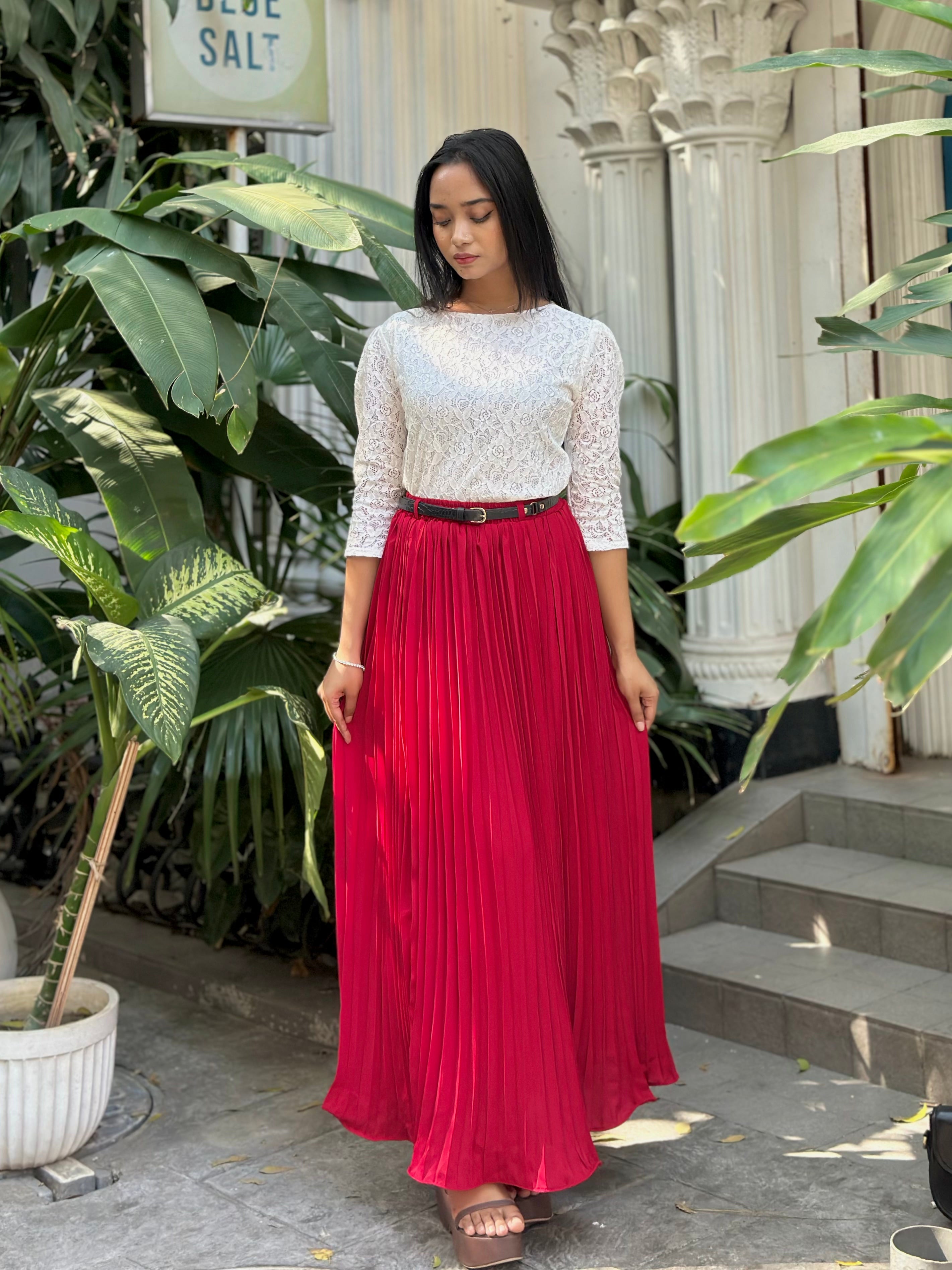 Woman wearing a white lace top and red pleated skirt standing outdoors with plants and steps in the background.