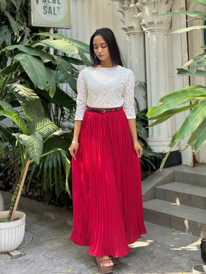 Woman wearing a white lace top and red pleated skirt standing outdoors with plants and steps in the background.