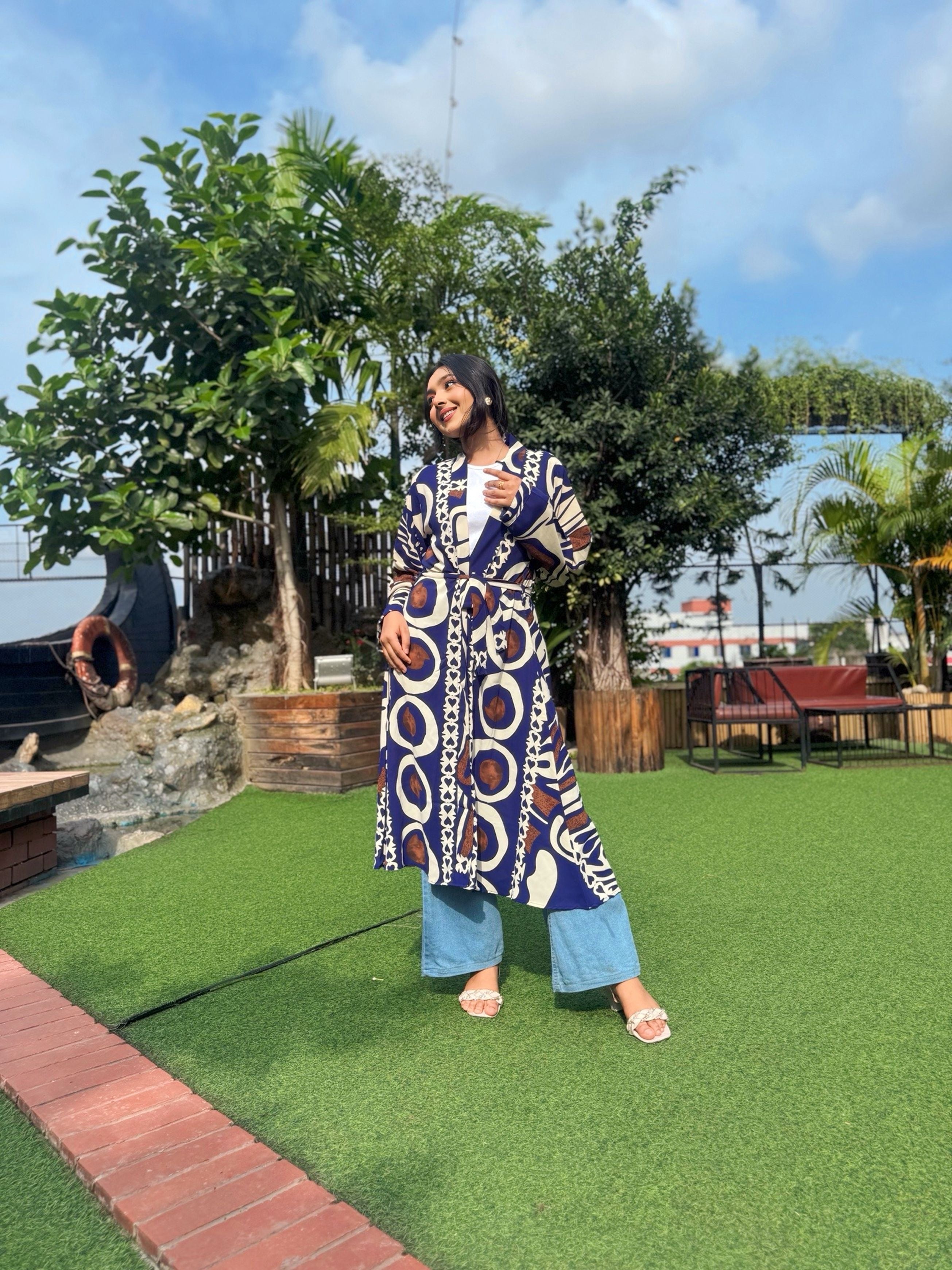 Woman wearing a long patterned shrug-style jacket and jeans with a handbag outdoors.