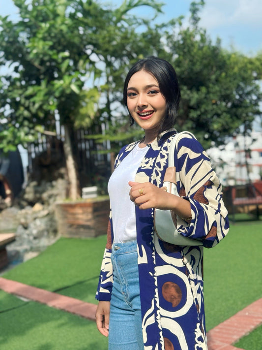 Woman wearing a long patterned shrug-style jacket and jeans with a handbag outdoors.