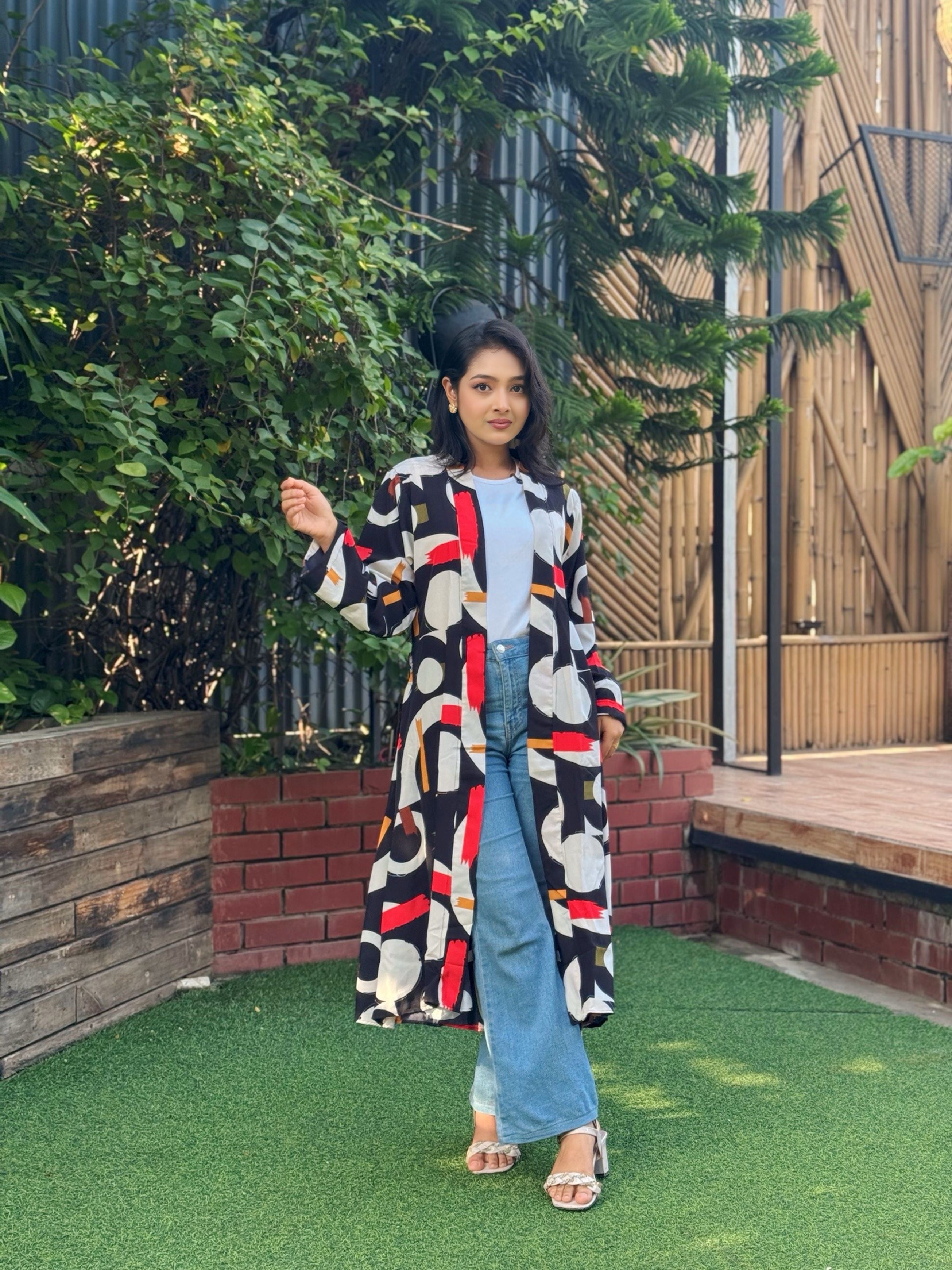 Woman wearing a colorful long shrug in an outdoor setting with plants and wooden structures.