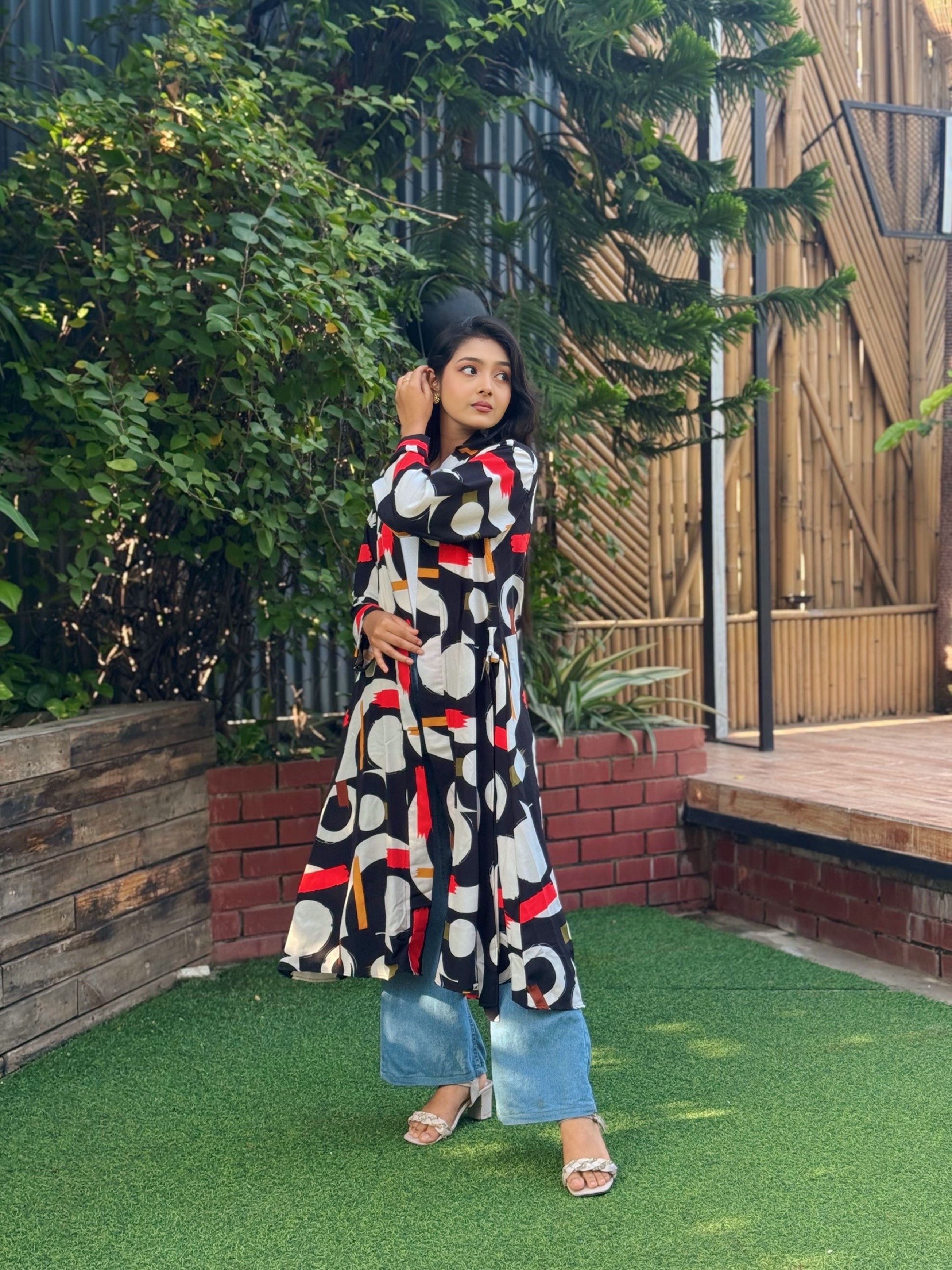 Woman in a patterned long shrug standing outdoors with greenery and wooden structures in the background