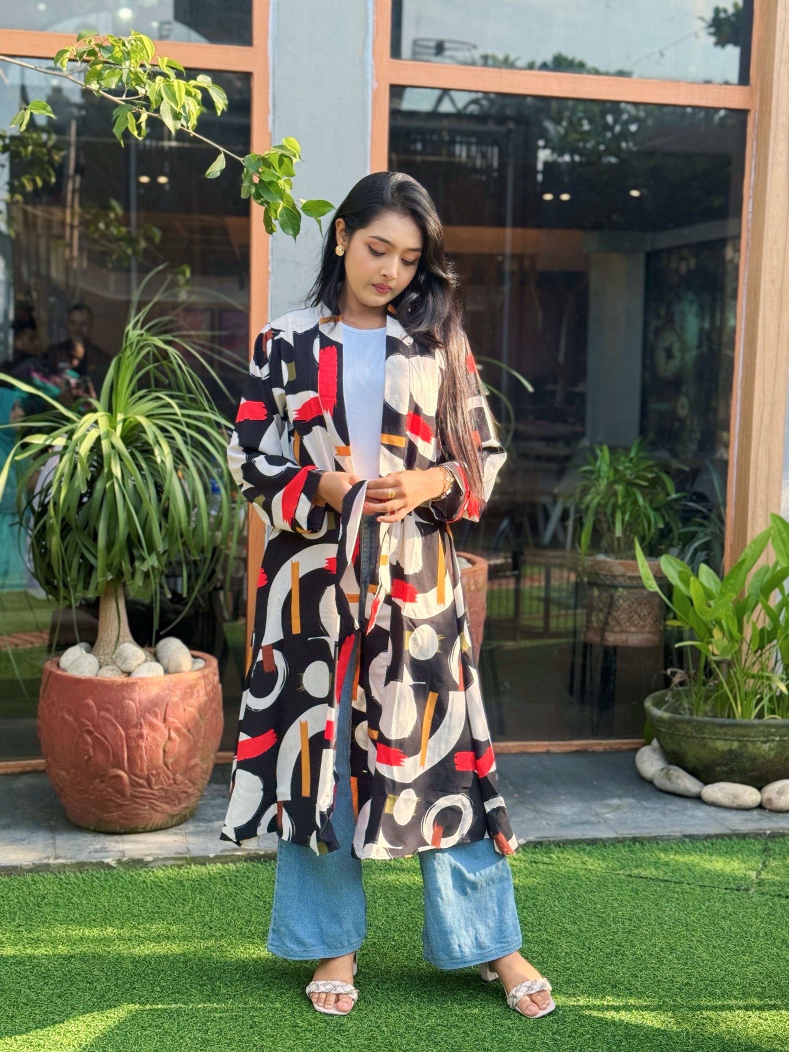 Woman wearing a patterned long shrug standing outdoors with plants and a building in the background