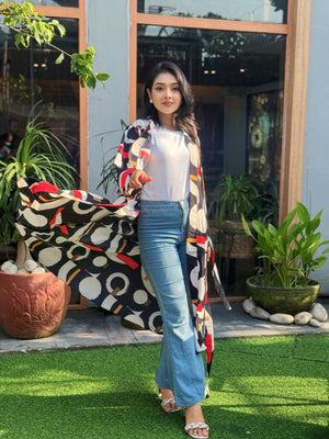 Woman standing outdoors with a colorful patterned long shrug, surrounded by plants.