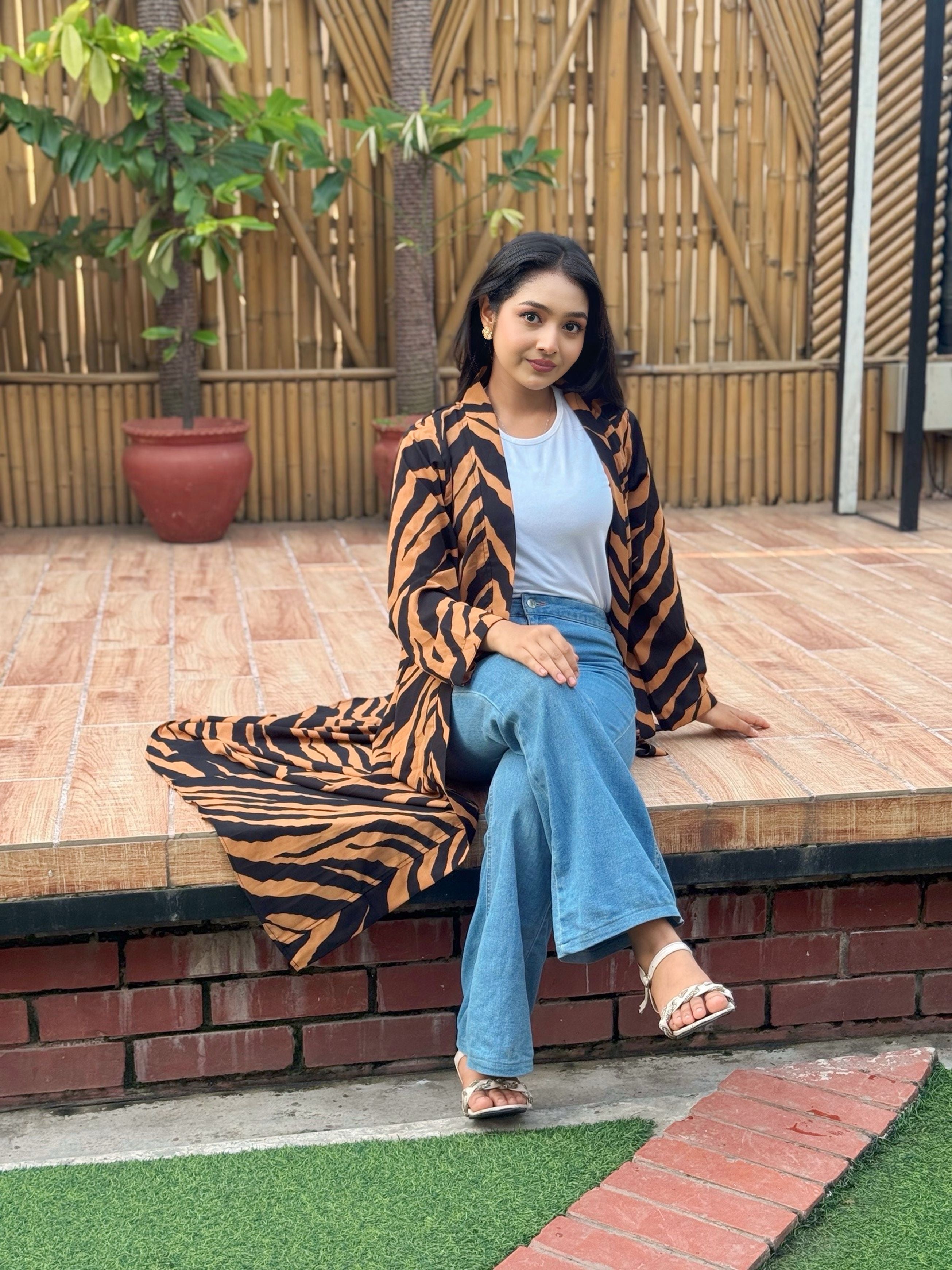 Woman sitting outdoors wearing a tiger-striped long shrug, light blue jeans, and white sandals.