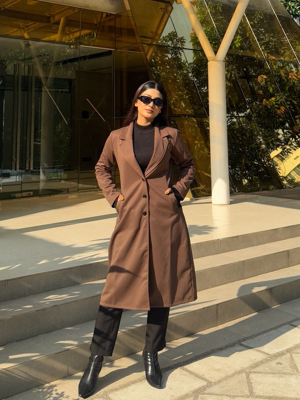 Person in a brown coat standing in front of a modern building with glass walls.