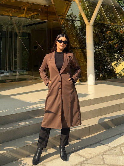 Person in a brown coat standing in front of a modern building with glass walls.