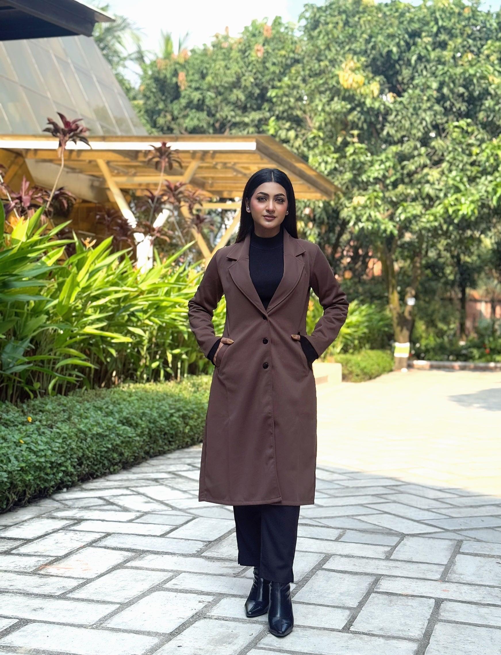 Woman in a brown coat standing on a paved path with greenery in the background