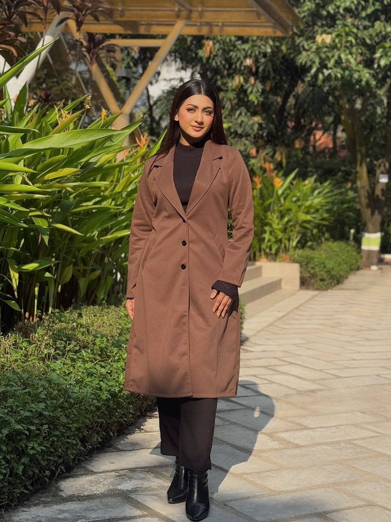Woman in a brown coat standing in a garden setting with greenery and a wooden structure.
