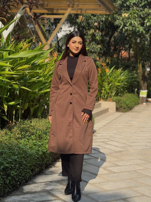 Woman in a brown coat standing in a garden setting with greenery and a wooden structure.